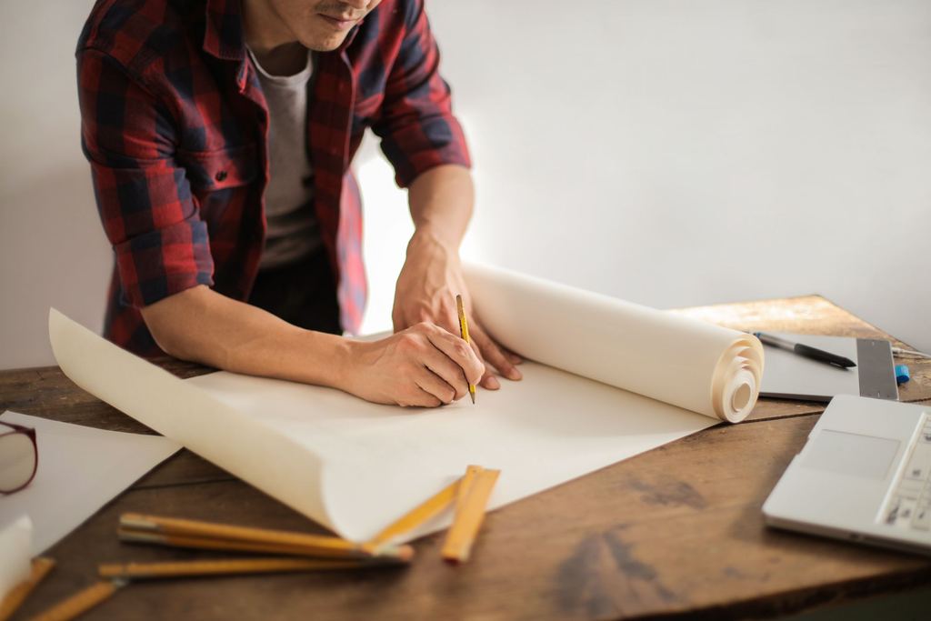 Person unrolling large printed plans across a desk beside drawing tools
